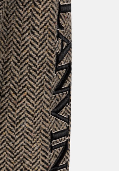 Tessuto di lana a motivo pied de poule con un grande logo ricamato nero sul lato. La texture è morbida e calda, caratterizzata da una palette di colori neutri.