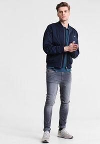 Marineblaues Bomberjackett mit Reißverschluss, gerippten Manschetten, über einem tealfarbenen Hemd getragen. Kombiniert mit grauen Slim-Fit-Jeans und hellgrauen Sneakers.