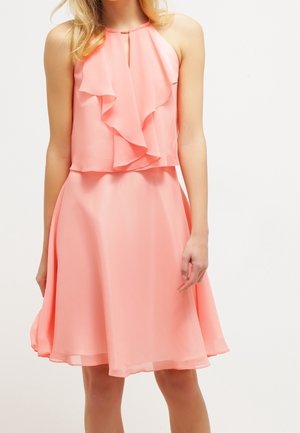 Femme portant une robe rose corail sans manches avec un détail en volant sur la poitrine et une jupe évasée, debout devant un fond uni.