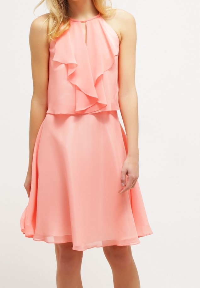 Femme portant une robe rose corail sans manches avec un détail en volant sur la poitrine et une jupe évasée, debout devant un fond uni.
