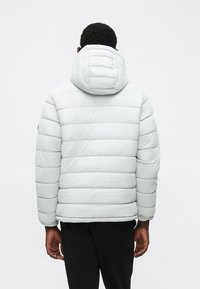 Ανοιχτό γκρι μπουφάν με κουκούλα, με επενδεδυμένα τμήματα, ελαστικούς μανσέτες και λεία υφή. Η πίσω πλευρά δείχνει χαλαρή εφαρμογή.