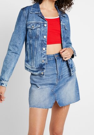 Vrouw draagt een blauwe spijkerjas en bijpassende spijkerrok over een rood cropped topje tegen een effen achtergrond.