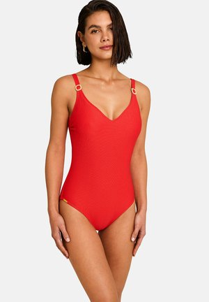 Vrouw met kort donker haar die een aansluitende rode badpak draagt met dunne schouderbandjes en gouden ringdetails op de bandjes.