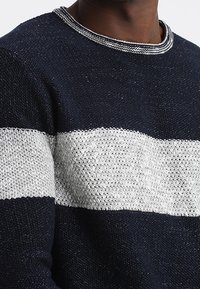 Pull en maille bleu marine et blanc avec un motif texturé, comportant une large bande horizontale blanche et un col rond avec une bordure contrastante.