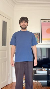 Άνδρας με γενειάδα και σγουρά μαλλιά, ντυμένος με μπλε κοντομάνικο και καφέ παντελόνι, στέκεται σε δωμάτιο με ξύλινο πάτωμα, τηλεόραση και κορνιζαρισμένη τοιχογραφία πίσω του.