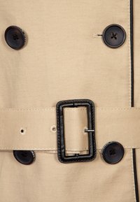 Beige trenchcoat detail met zwarte knopen, een zwarte riemgesp en een textiel met een contrasterende steek langs de randen.