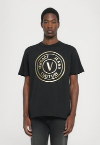 Tricou din bumbac negru, cu un logo circular mare de culoare aurie, având textul "VERSACE JEANS COUTURE" și litera centrală "V". Mâneci scurte, guler rotund.