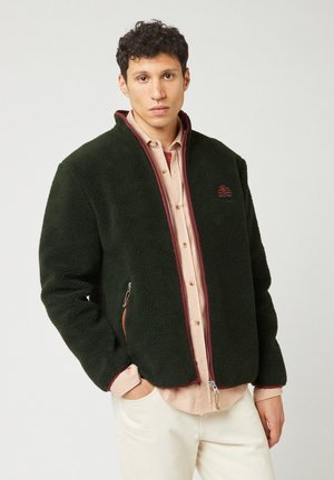 Green fleece jacket with a high collar, burgundy zippers, and side pockets. Worn over a light striped shirt. Textured, warm fabric.