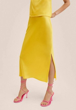 Femme portant une jupe midi jaune avec une fente latérale et des sandales à talons hauts roses à lanières, debout avec une main sur la hanche.