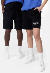 Černé bavlněné šortky s elastickým pasem, kapsami a bílým logem KANI. Vybavené bílými teniskami a ponožkami. Jednoduchý design.