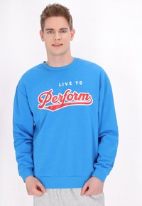Kék pamut pulóver, hosszú ujjal és kerek nyakkivágással. A középen elhelyezett piros és fehér szöveg így olvasható: "LIVE TO Perform."
