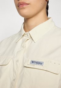 Camicia button-up beige chiaro con colletto, dotata di due tasche frontali e etichetta Columbia PFG. Il tessuto ha un aspetto leggermente testurizzato.