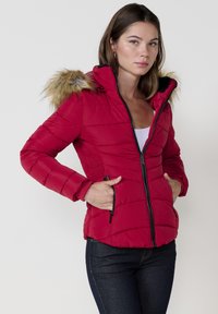 Raudona pūkuota striukė su kailiu išklota kapuce, užtrauktuko priekine dalimi, šoninėmis kišenėmis ir tekstūruotu audiniu šonuose.