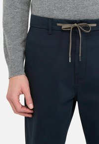 Pantalon bleu marine à la texture lisse, doté d'une taille élastiquée avec un cordon de serrage beige contrastant et d'accents métalliques élégants.