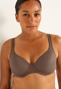 Brun, vadderad behå med justerbara band och slät textur. Har en stödjande design och enkel sömnad längs kanterna.