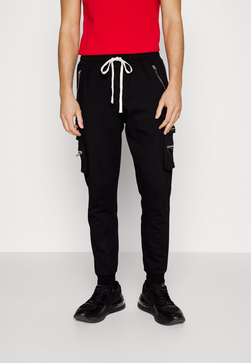 Man med svarta cargo-joggingbyxor med vit dragsko och svarta sneakers, stående mot en enkel vit bakgrund.
