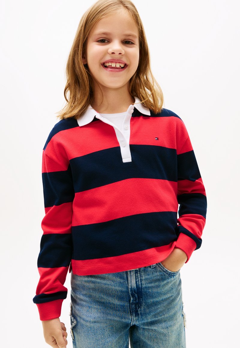 Μακρυμάνικο πόλο μπλουζάκι με κόκκινες και ναυτικές ρίγες, λευκό γιακά και πατιλέτα, συνδυασμένο με ανοιχτόχρωμα, φθαρμένα τζιν παντελόνια.