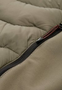 Stworzona z pikowanego oliwkowego materiału, z teksturowaną beżową sekcja, z czarnym zamkiem błyskawicznym z czerwonymi akcentami do zapięcia.