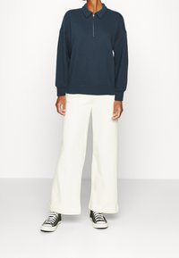 Felpa blu navy con zip e collo, vestibilità rilassata. Abbinata a pantaloni bianchi a gamba larga e sneakers nere. Texture liscia, design minimale.