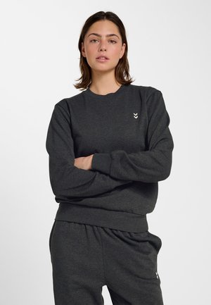 Junge Frau trägt dunkelgraues Sweatshirt und passende Jogginghose mit kleinen weißen Chevron-Logos, steht mit verschränkten Armen.