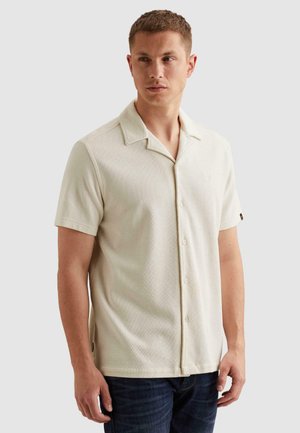 Mann trägt kurzärmliges, cremefarbenes Hemd mit Knöpfen und Kragen, steht vor hellem einfarbigem Hintergrund und schaut zur Seite.