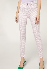 Mujer vestida con pantalones de cuadros en tonos rosa claro y blanco de corte slim y zapatos de tacón verde tipo slingback, de pie frente a un fondo liso.