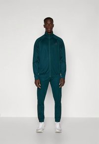 Τσάιλ φόρμα από απαλό ύφασμα, με μπουφάν με φερμουάρ και ψηλό γιακά, καθώς και αντίστοιχο στενό παντελόνι με πλαϊνές ρίγες.