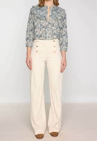 Blus med blommönster och volangkrage, knäppning framtill och trekvartslånga ärmar, kombinerad med högmidjade krämfärgade byxor med guldknappar.