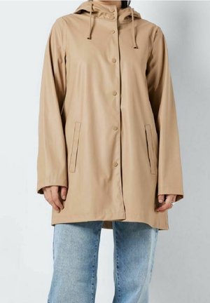 Persona que lleva un impermeable beige abotonado con capucha y dos bolsillos delanteros, combinado con vaqueros azul claro sobre un fondo liso.