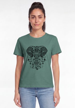 Frau trägt grünes T-Shirt mit detailreichem schwarzem Elefantendesign und dem Text "Be Strong", kombiniert mit hellblauen Jeans, steht vor weißem Hintergrund.