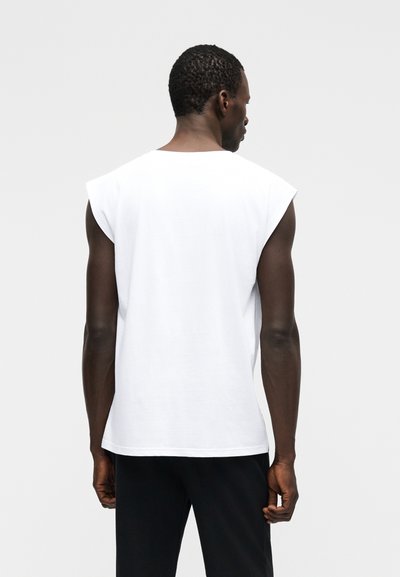 Άνδρας με κοντά σγουρά μαλλιά, ντυμένος με λευκό αμάνικο πουκάμισο και μαύρο παντελόνι, στέκεται με την πλάτη του στραμμένη προς ένα απλό φωτεινό φόντο.