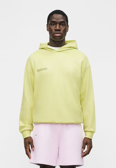 Νεαρός άνδρας στέκεται αντιμετωπίζοντας προς τα εμπρός φορώντας ανοιχτό κίτρινο φούτερ με μικρό κείμενο στο στήθος και ανοιχτό ροζ σορτς, πάνω σε απλό φόντο.