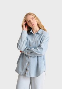 Lichtblauwe denimshirt met knoopsluiting, lange mouwen en twee borstzakken. Heeft een relaxte pasvorm en een gebogen zoom.