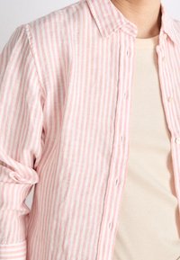 Jasnoróżowa i biała w paski koszula zapinana na guziki z kołnierzykiem, wykonana z lnu. Posiada podwinięte rękawy i widoczną beżową koszulę wewnętrzną.