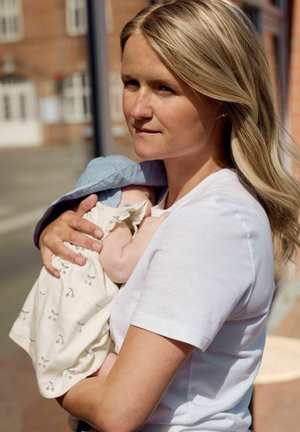 Femme aux cheveux longs blonds portant un T-shirt blanc tenant un bébé enveloppé dans une robe claire à motif de cerises, en plein air près de bâtiments.