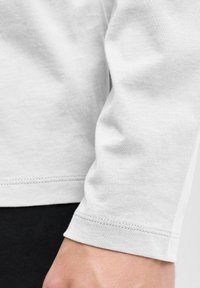 Camisa blanca de manga larga con una textura de algodón suave, con un dobladillo cosido en el puño y un diseño elegante. Mano parcialmente visible.