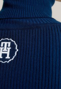 Close-up van marineblauwe geribbelde gebreide stof met wit geborduurd "TH"-logo binnen een gegolfde cirkel.