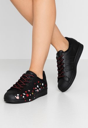 Jambes portant des baskets noires avec des motifs de cœurs rouges et blancs et des lacets noirs assortis avec de petits cœurs rouges sur un sol clair.
