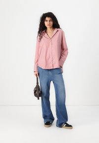 Camisa de botão com riscas vermelhas e brancas, com um corte relaxado, combinada com jeans de perna larga azul claro e ténis pretos. Segurando uma pequena bolsa preta.