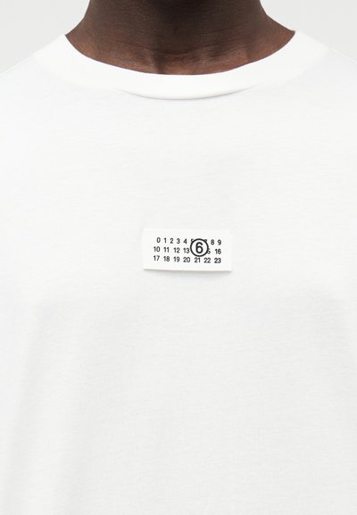 Biele bavlnené tričko s okrúhlym výstrihom a malým obdĺžnikovým štítkom. Štítok zobrazuje čierne čísla a kruhový detail.