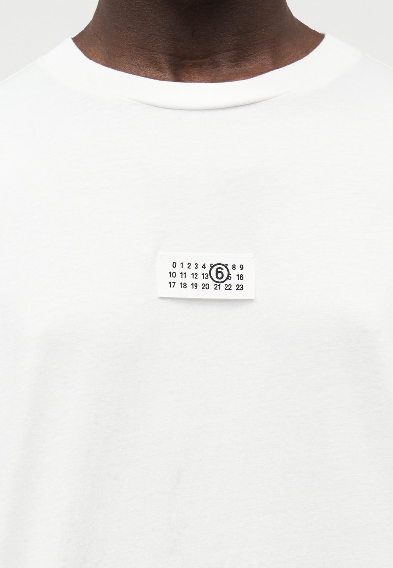 Hvid t-shirt i bomuld med rund halsudskæring, der har en lille rektangulær mærkat. Mærkaten viser sorte tal og en cirkulær detalje.