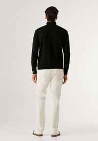 Maglione nero a maglia con colletto alto, abbinato a jeans in denim bianco. L'outfit presenta una vestibilità slim ed è ambientato su uno sfondo neutro.