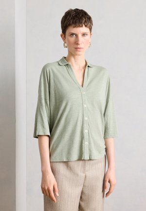Femme aux cheveux courts et bruns porte un chemisier vert clair boutonné avec des manches aux coudes et un pantalon beige à fines rayures, debout contre un mur gris.