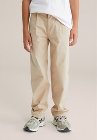 Bežīgas kokvilnas bikses ar brīvu piegriezumu, ar priekšējiem kabatām un gludu tekstūru, apvienotas ar pelēkiem un melniem sporta apaviem.