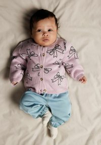 Lyserosa cardigan med svarte sløyfemønstre, gullknapper, kombinert med lyseblå bukser og hvite sokker, liggende på en beige overflate.