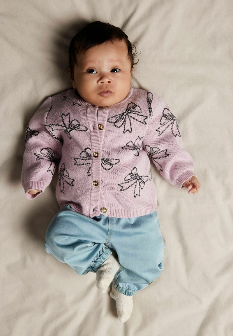 Lyserosa cardigan med svarte sløyfemønstre, gullknapper, kombinert med lyseblå bukser og hvite sokker, liggende på en beige overflate.