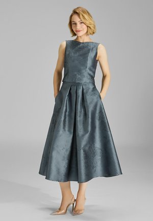 Ciemnozielona sukienka bez rękawów z dopasowanym topem i plisowaną spódnicą w kształcie litery A. Posiada subtelny wzór kwiatowy oraz boczne kieszenie. Przezroczyste, wysokie obcasy.