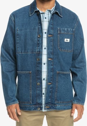 Mand iført en blå denimjakke over en lyseblå ternet skjorte og beige bukser, står med armene afslappet ned langs siderne.