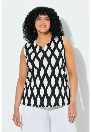 Kobieta o kręconych włosach uśmiechająca się, ubrana w bezrękawnik w czarno-białe wzory i białe spodnie, stojąca na jasnym tle.