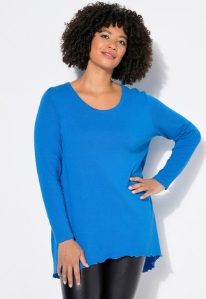 Tunica a maniche lunghe di un brillante blu con scollatura a barca e orlo irregolare. Realizzata in un morbido tessuto testurizzato. Abbinata a leggings neri.